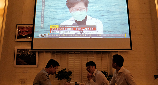 Friends have a drink as a projection is shown of a live press conference held by Chief Executive Carrie Lam, wearing a face mask as a precautionary measure against the COVID-19 coronavirus, in Hong Kong on March 23, 2020. Anthony WALLACE / AFP